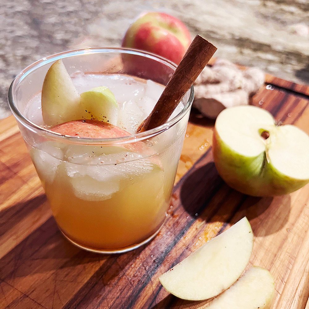 Apple Spice Gin Sour pictured on a wooden cutting board surrounded by cut apples and fresh ginger, with a cinnamon stick.